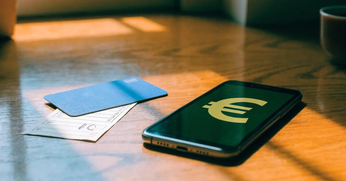 Ein Smartphone liegt mit dem Display nach unten auf einem Holztisch, daneben ein paar Euro-Münzen und eine unbeschriftete Plastikkarte im Morgenlicht.