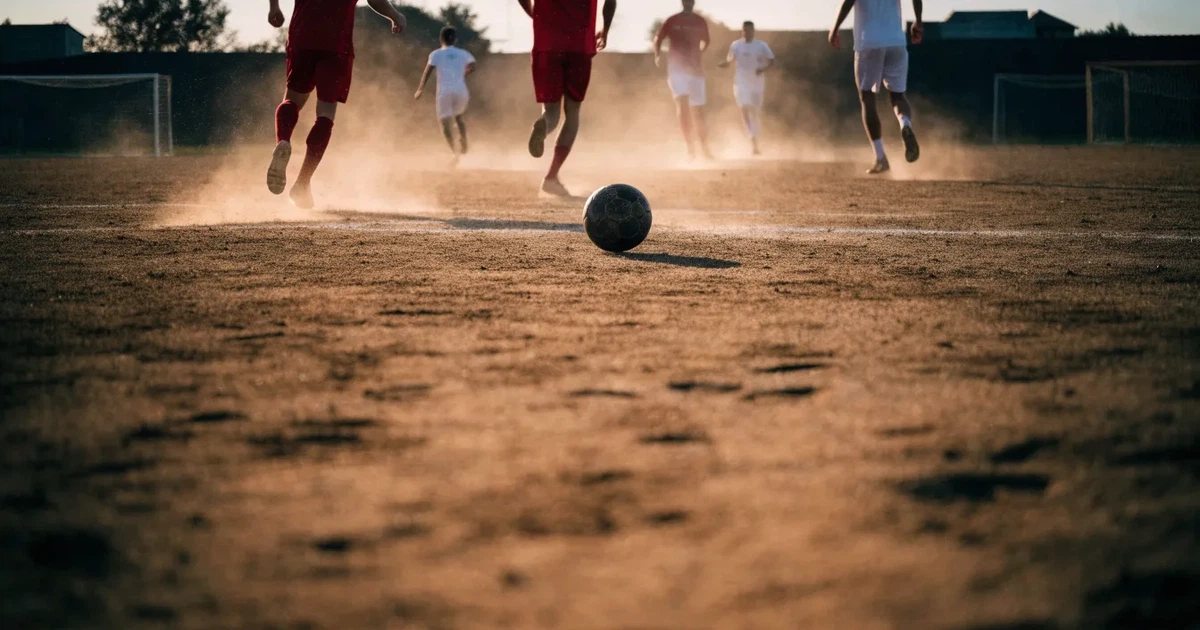 Ein sandiger Fußballplatz am Rand einer Stadt in warmem Abendlicht, Staub in der Luft und zwei Tore in der Ferne.