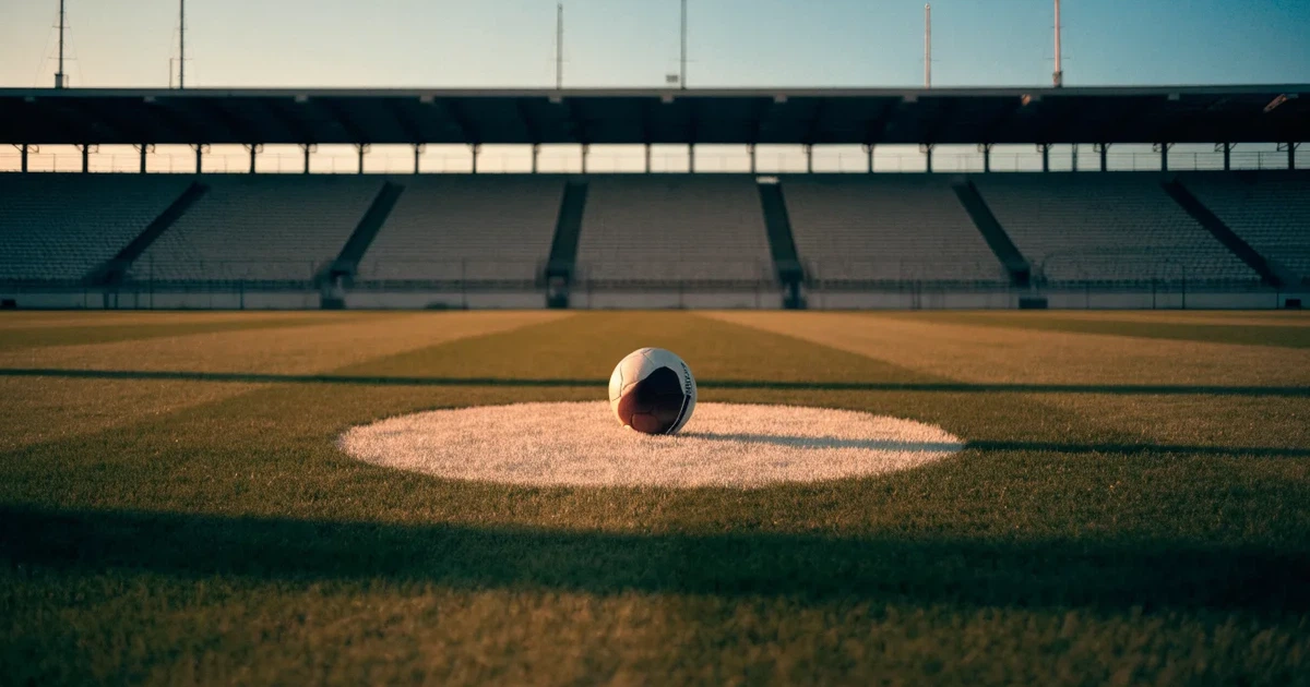 Ein Fußball liegt auf dem Anstoßpunkt eines leeren Rasenplatzes, im Hintergrund eine große Stadiontribüne im Abendlicht.