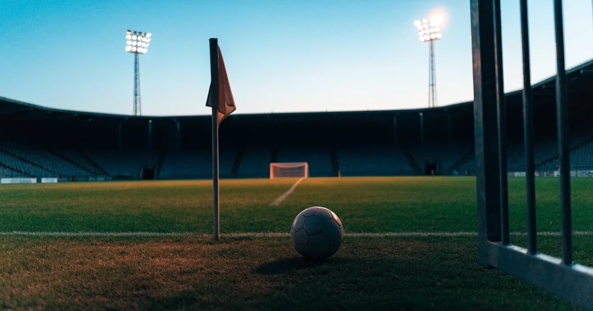Blick durch ein Stadiontor auf einen grünen Rasen im Abendlicht, ein Ball ruht an der Eckfahne, die Tribünen sind leer.