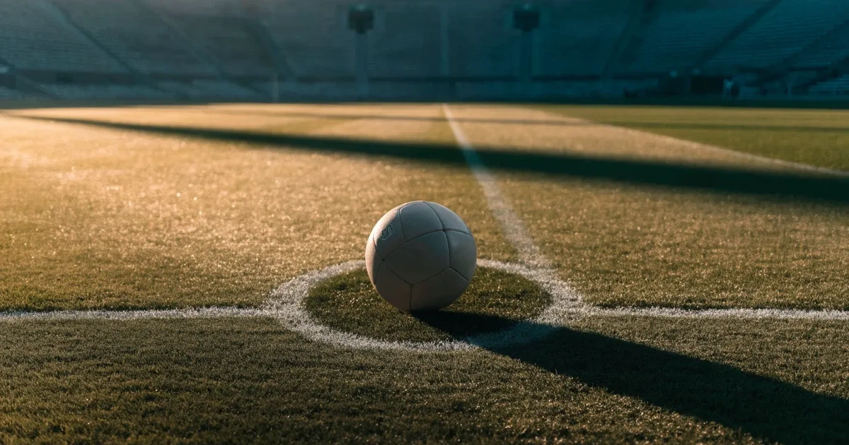Ein Fußball liegt auf dem Mittelkreis eines Stadionrasens am frühen Morgen, im Hintergrund leere Ränge ohne erkennbare Beschriftung.