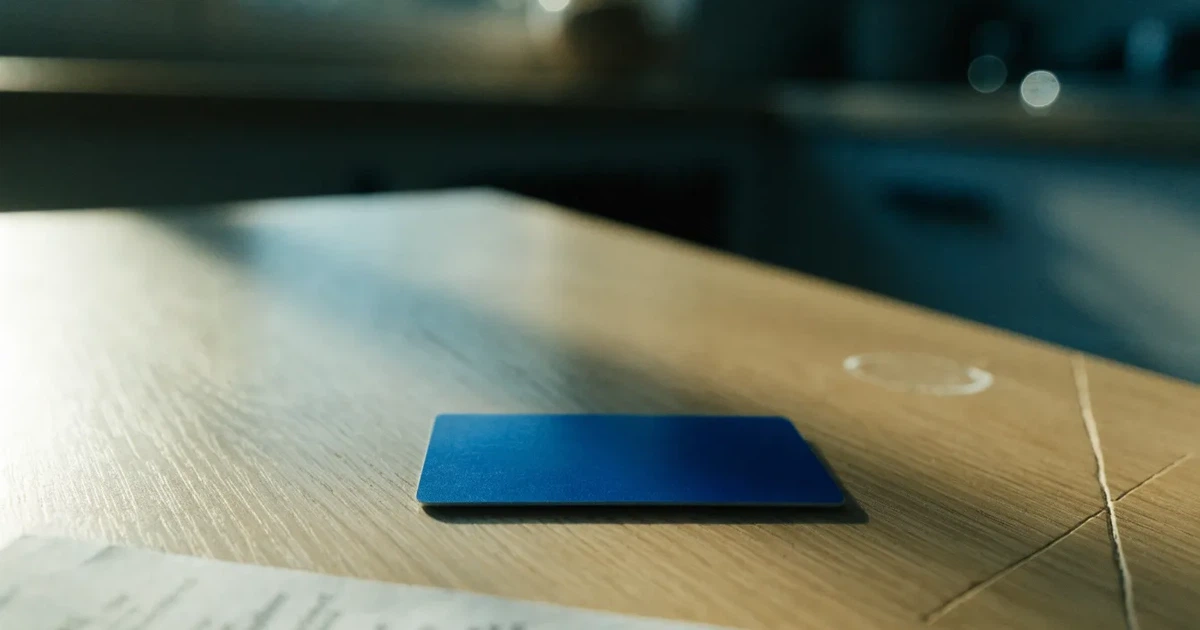 Eine einfache Plastikkarte liegt ohne erkennbare Aufdrucke neben einem kleinen Stapel Münzen auf einem hellen Holztisch im Morgenlicht.