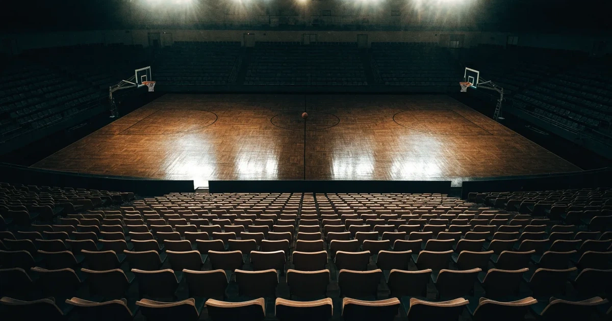 Eine leere Basketballhalle von einer oberen Tribünenreihe aus gesehen, Parkett und Körbe im Licht der Hallenstrahler.