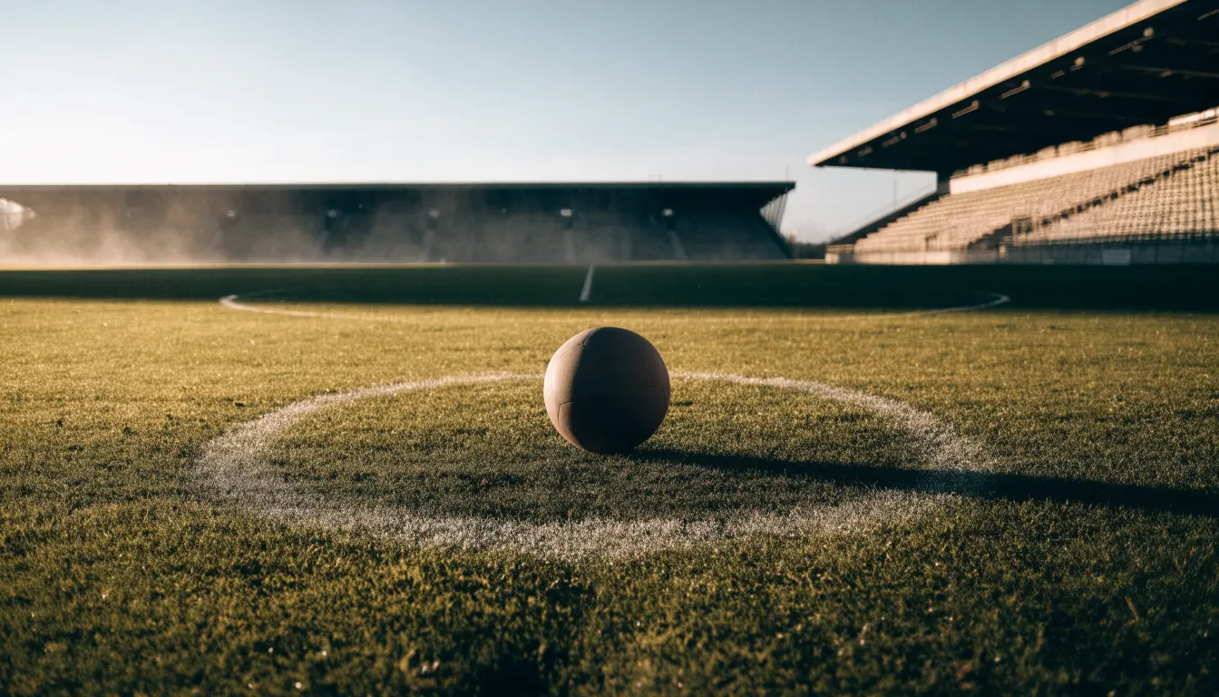 Ein Fußball liegt auf dem Anstoßpunkt eines leeren Rasenplatzes im frühen Morgenlicht, dahinter eine leere Tribüne.