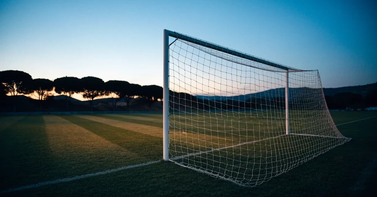 Ein Fußballtor mit Netz auf einem leeren Platz im Abendlicht, der Rasen frisch gemäht, im Hintergrund Zypressen.