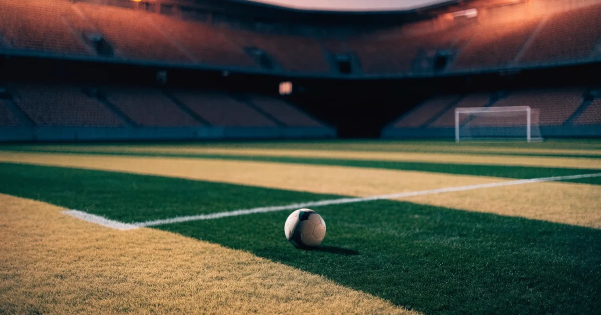 Ein Fußballplatz mit frisch gemähtem Rasen im Abendlicht, ein Ball liegt im Strafraum, im Hintergrund unscharf eine leere Tribüne.