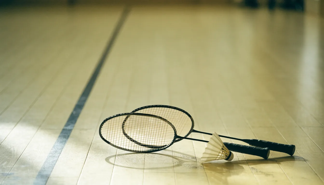 Zwei Badmintonschläger und ein Federball liegen auf einem Holzboden neben der Seitenlinie, weiches Licht fällt von der Seite ein.