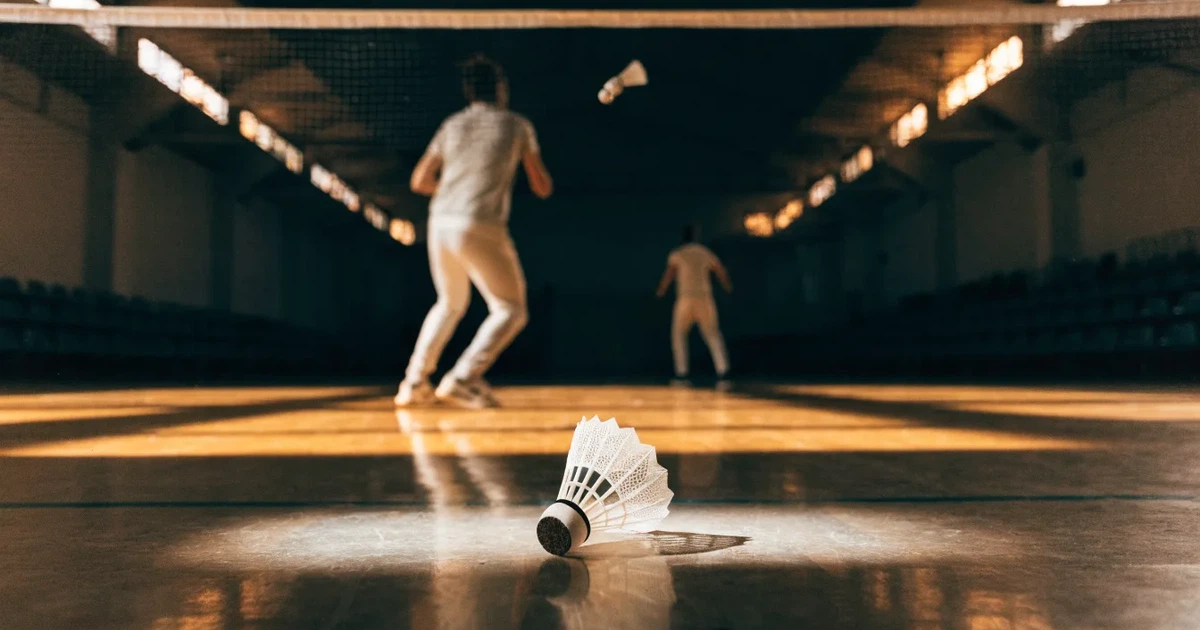 Eine leere Badmintonhalle mit gespanntem Netz und hellen Hallenlichtern, ein Federball liegt auf dem Hallenboden.