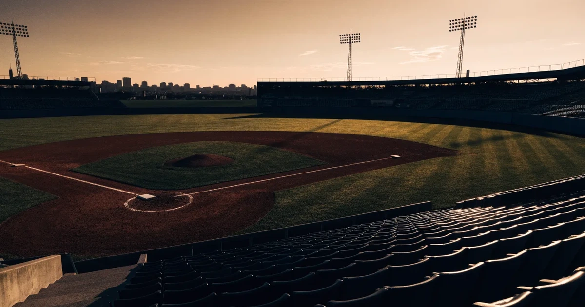Ein Baseballfeld mit dem hellbraunen Infield und gepflegtem Rasen, von der Tribüne aus gesehen, die Ränge leer, tiefe Abendsonne.