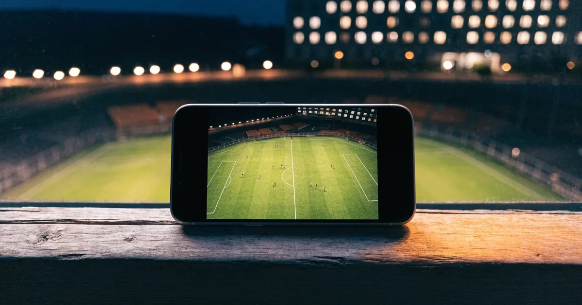 Ein Fußballstadion bei Flutlicht aus der Ferne gesehen, davor eine Straße und leuchtende Fenster eines Nebengebäudes.
