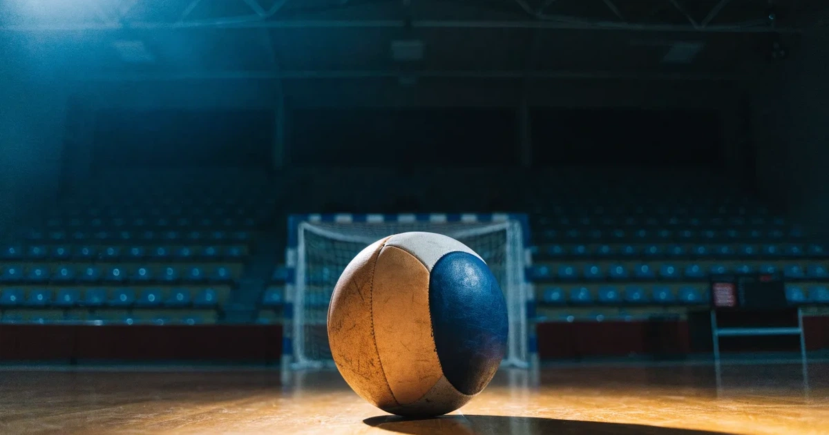 Ein Handball liegt auf dem Hallenboden vor einem Tor mit Netz, die leere Halle wird von Flutlicht beleuchtet.