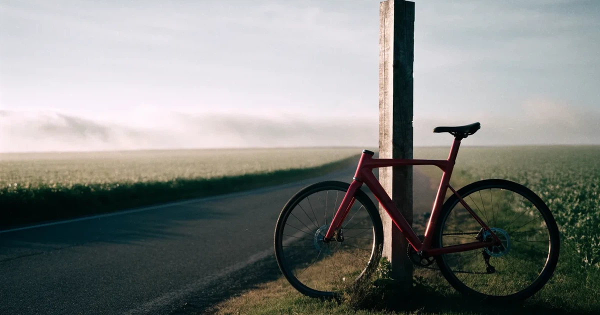Eine Landstraße zwischen Feldern im Morgenlicht, am Straßenrand lehnt ein Rennrad ohne Aufschrift an einem Pfosten.
