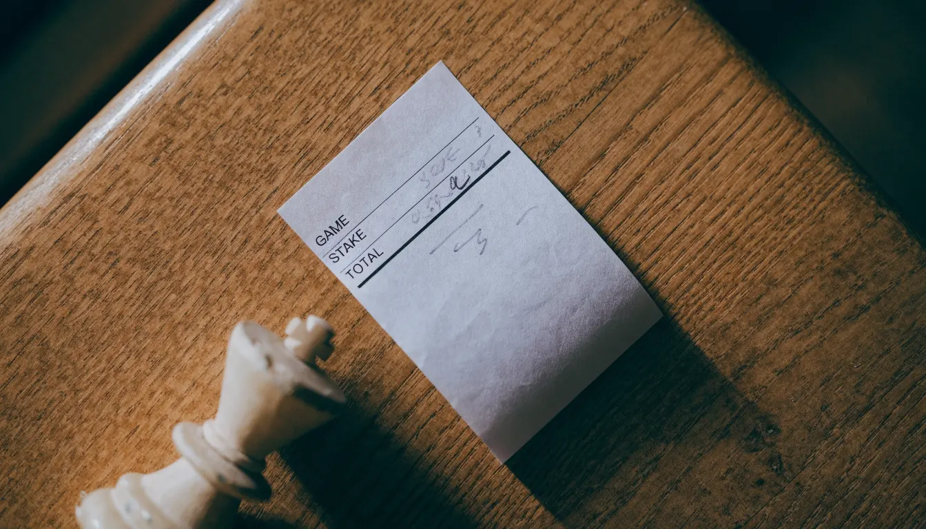Ein leerer Wettschein aus Papier liegt neben einem umgekippten weißen Schachkönig auf einem Holztisch.