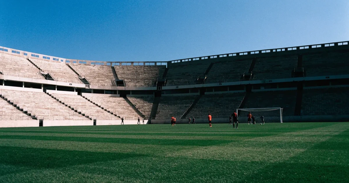 Ein Fußballstadion mit hellen Rängen unter strahlend blauem Himmel, davor ein sonnenbeschienener Rasen mit frisch gemähten Streifen.