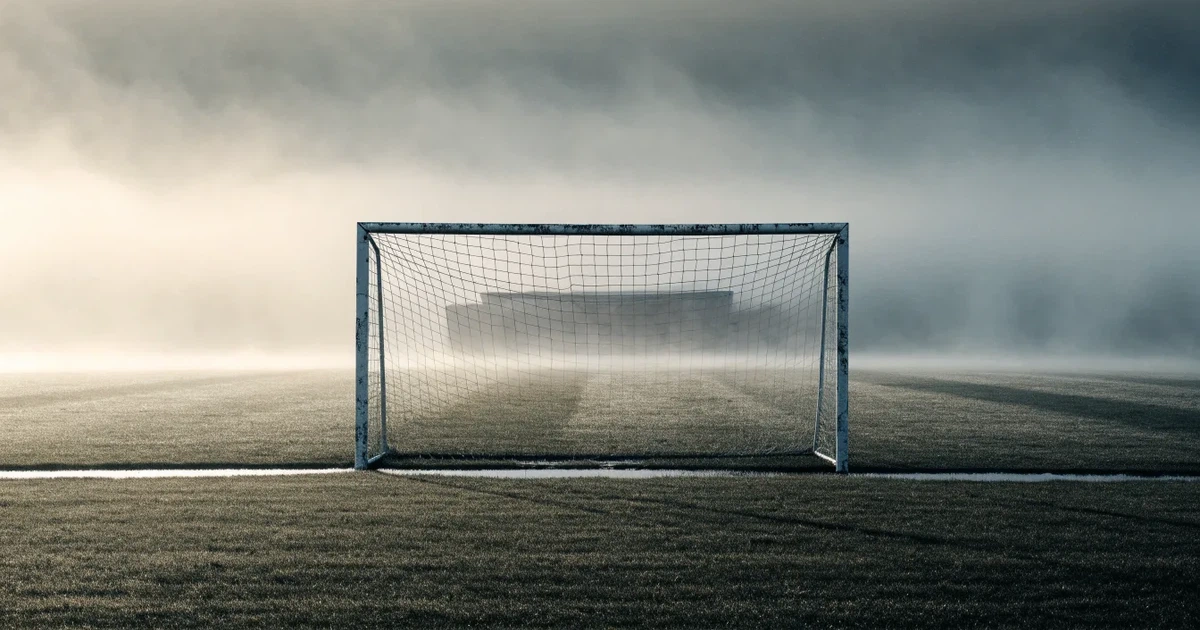 Ein Fußballtor mit Netz steht im Morgennebel auf einem leeren Rasenplatz, im Hintergrund die Silhouette einer kleinen Tribüne.