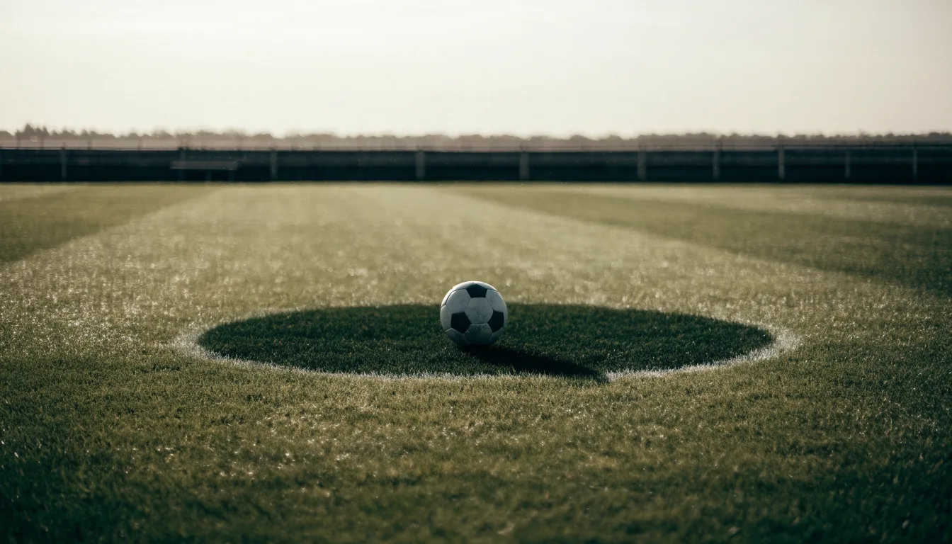 Ein Fußball liegt allein auf dem Anstoßpunkt eines leeren Rasenplatzes im Morgenlicht, am Rand ein niedriger Zaun.