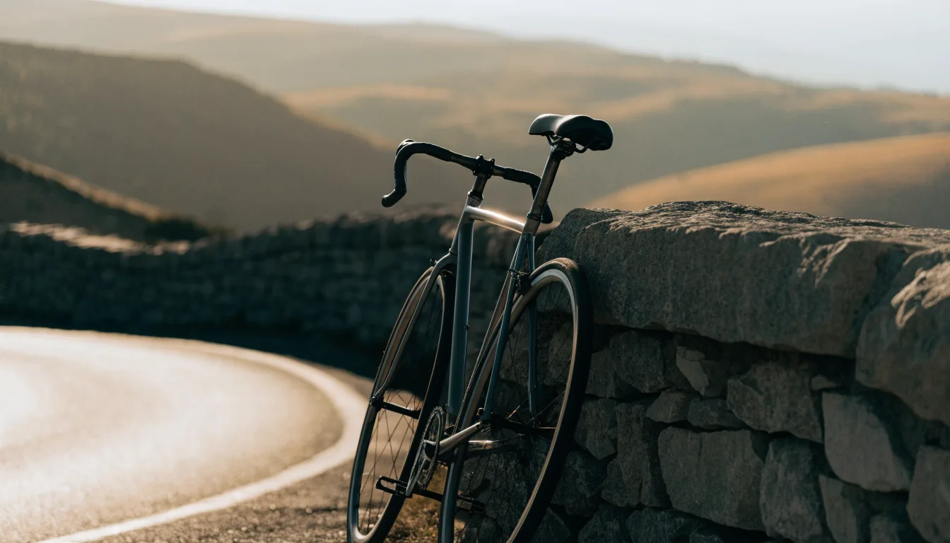 Ein Rennrad lehnt an einer Steinmauer in einer Bergkurve, im Hintergrund eine weite Hügellandschaft im Gegenlicht.