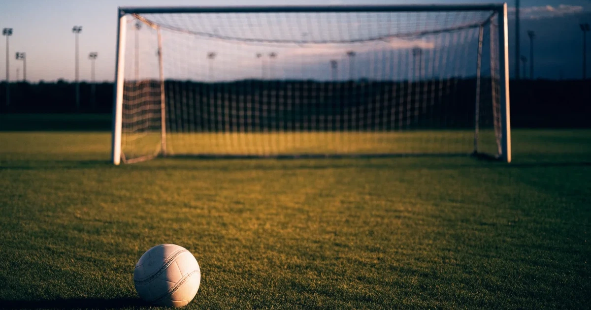 Ein Fußballtor mit Netz im Gegenlicht auf einem leeren Platz am Abend, davor ein Ball auf dem Rasen.