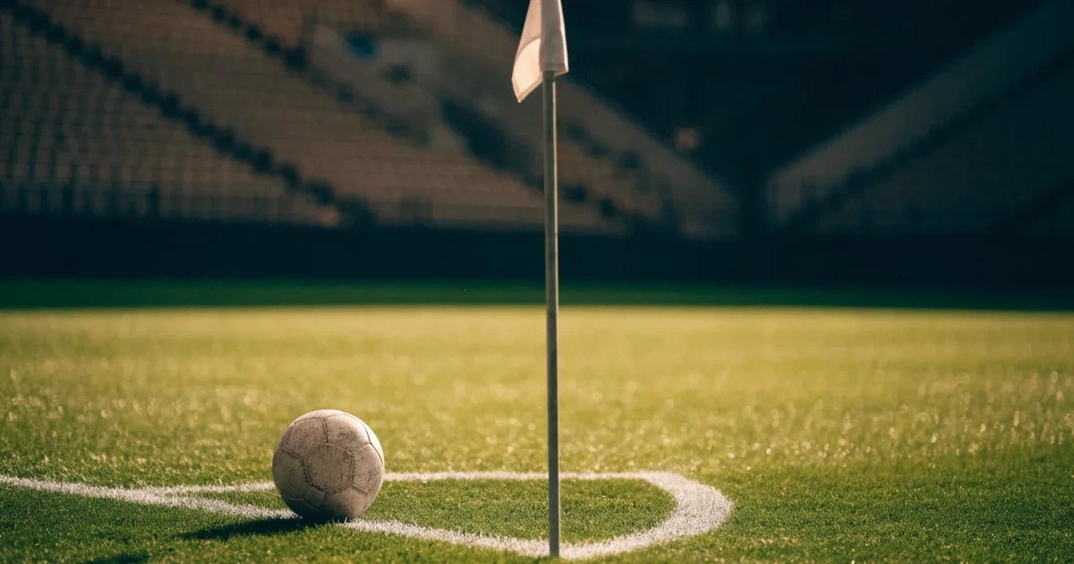 Ein Fußball liegt am Viertelkreis an der Eckfahne eines Rasenplatzes, im Hintergrund leere Tribünen in warmem Abendlicht.